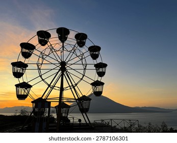 Nostalgic twilight view of an empty amusement park Ferris wheel silhouetted against a vibrant orange sunset sky, with a mountain and sea backdrop in Lembata Island, Indonesia.