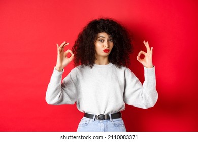 Not Bad. Impressed Curly Woman Nod In Approval, Pucker Lips And Showing Okay Signs, Like Good Thing, Standing Satisfied On Red Background