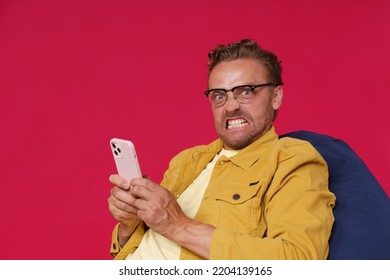 Not Happy, Annoyed Handsome Man In Glasses Hold Smartphone In Hands Looking At Camera With Irritated Look Wearing Yellow Denim Jacket Sitting On Bag Chair Isolated On Red Background. 