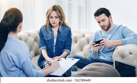 Not Interested. Nice Handsome Indifferent Man Sitting On The Sofa And Playing On His Smartphone While Being Not Interested It The Psychological Therapy