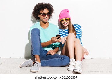 Not noticing anyone else. Funky young couple smiling and looking at the mobile phone while sitting against white background