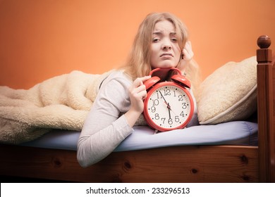 Not Want To Wake Up Concept. Funny Young Woman In Bed Waking Up Late. Unhappy Sleepy Girl Holds Red Alarm Clock.