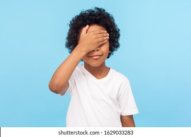 I'm not watching. Portrait of cute little boy with curls covering eyes with hand not to look at something forbidden, hiding eyes playing peek a boo. indoor studio shot isolated on blue background