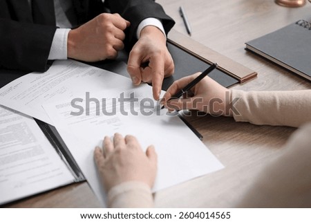 Notary pointing at document and client putting signature at wooden table indoors, closeup