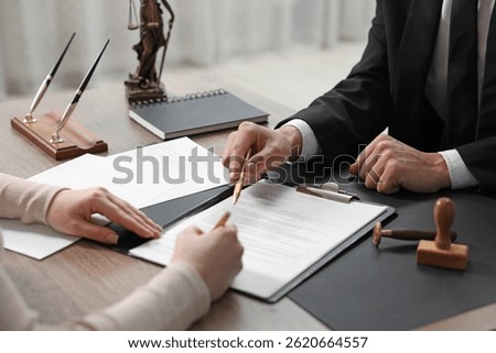 Notary pointing at document and client putting signature at wooden table indoors, closeup