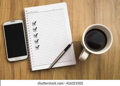 Note Book With Five Check Marks, Smartphone, A Pen And A Cup Of Coffee On Wooden Background, Copy Space. Background For Anything.