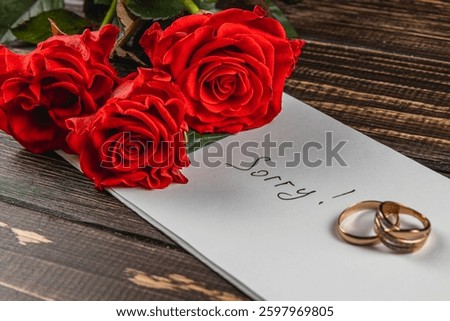 A note saying sorry is placed on a wooden surface alongside three red roses and two wedding rings, symbolizing sadness and remembrance of a love lost.
