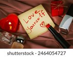 A note to self with call to love yourself drawing with red lipstick on a silk cloth flatly. A burning heart-shaped red candle, gift box Valentines Day