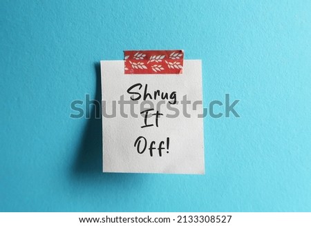 Note stick on blue wall with handwritten text SHRUG IT OFF, concept of to treat something as if it is not important or not a problem, to ignore someone or something unpleasant  