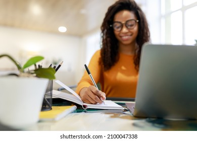 Note Taking Concept. Smiling Young Black Woman In Glasses Sitting At Desk Working On Laptop And Writing Letter In Paper Notebook, Selective Focus On Hand Holding Pen. Happy Millennial Female Using Pc