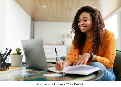 Note Taking Concept. Smiling young African American woman sitting at desk working on laptop and writing letter in paper notebook, holding pen and looking at screen. Happy millennial female using pc - Shutterstock ID 2122956533