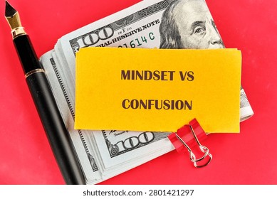 A note with the words mindset vs confusion sits on top of a stack of cash alongside a pen and a paperclip. The scene shows a workspace setup with financial elements.