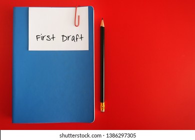 A Note Written FIRST DRAFT Clip With A Blue Notebook, With A Pencil, On Red Background. Concept Of Finishing First Draft On Book Writing Or Business Plan Or Creative Project