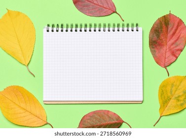 A notebook with a checkered pattern among yellow and red cherry leaves on a green background, top view