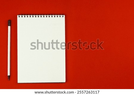 A notebook with an even spring at the top with white pages and a white pencil with a gray lead and a black eraser lie on a red background on the left with space for text. High quality photo