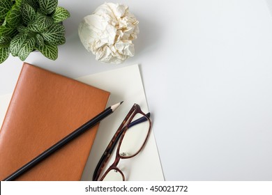 Notebook, Glasses, Pencil, Crumpled Ball And Plant Potted On White Desk Background