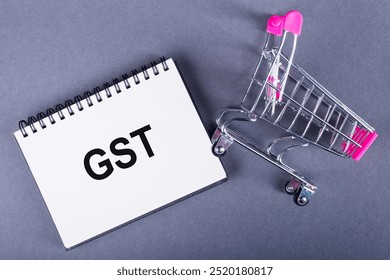 A notebook with GST lettering next to a miniature shopping cart, representing the concept of Goods and Service Tax. - Powered by Shutterstock