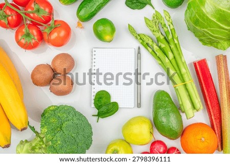 Notebook for meal planning surrounded by fresh fruits and vegetables including broccoli, asparagus, avocado, banana and tomatoes.