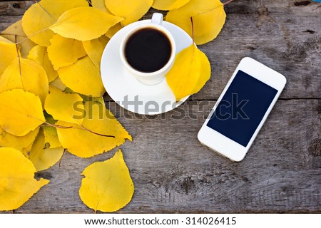 Notebook, mobile phone, cup of coffee and yellow autumn leaves on the old wooden table.