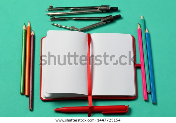 Notebook for\
notes in the red cover. With a red bookmark and pen. Lies opened on\
a mint background. Beside a set of colored pencils and accessories\
for drawing. School\
supplies.