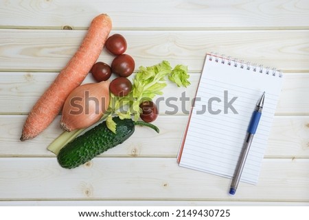 Notebook, pen and different vegetables from the regular diet on the background of light wooden boards. Vegetarian shopping. Budget planning, making a shopping list before going to the store.