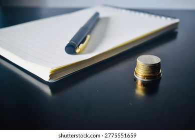 A notebook and pen placed next to a stack of coins on a dark surface, symbolizing financial goal setting and accounting.