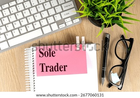 Notebook with pink note labeled sole trader on a wooden desk with keyboard, pen, glasses and green plant . 