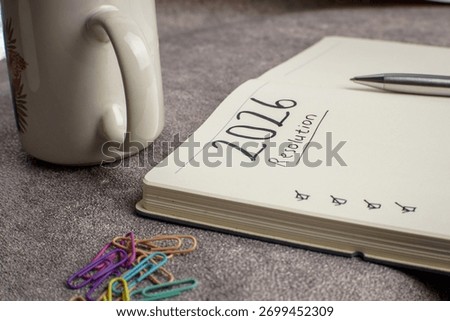 A notebook with “2026 Resolution” text and checked boxes beside a glass, pen, paper clips, and small boxes on gray cement background, symbolizing new year goals and planning concept.