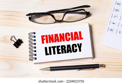 A notebook with the text FINANCIAL LITERACY, a pen and glasses lies on a wooden office table. Top view, flat lay. - Shutterstock ID 2234013735