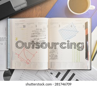 Notebook with text inside Outsourcing on table with coffee, some diagrams on paper and laptop 