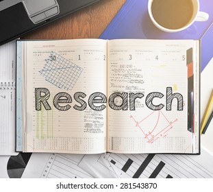Notebook with text inside Research on table with coffee, laptop and some sheet of papers with charts and diagrams