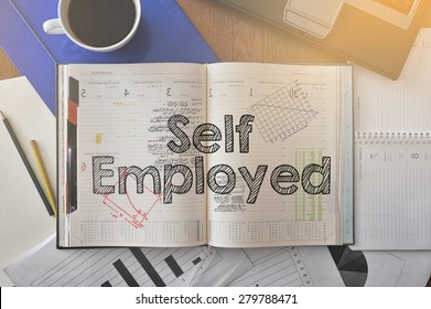 Notebook with text inside Self Employed on table with coffee, some diagrams on paper and laptop 