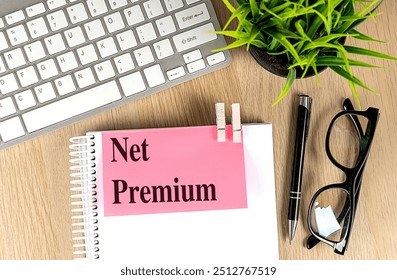  Notebook with the text net premium on a pink sticky note, next to keyboard, pen, glasses, and plant on a wooden desk . 