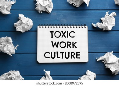 Notebook With Text Toxic Work Culture And Crumpled Paper On Blue Wooden Table, Flat Lay