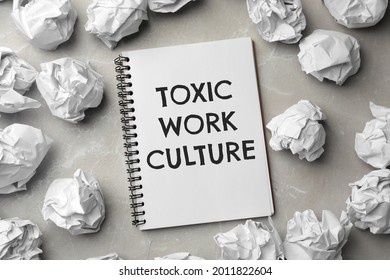 Notebook With Text Toxic Work Culture And Crumpled Paper On Grey Marble Table, Flat Lay