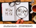 A notebook with the words "NEW YEAR" and "NEW DREAMS" written on it. A magnifying glass is held over the words "NEW DREAMS," highlighting their importance.