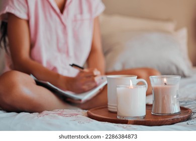 Notebook, writing and woman with home candles for healing journal, calm planning or self care inspiration in bedroom. Creative person or writer for mindfulness goals, meditation or peace notes in bed - Powered by Shutterstock