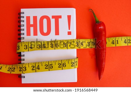 A notebook with a written text HOT, measuring tape and chilli on red background. Diet concept and scoville heat scale.
