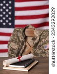 Notebooks and diploma on wooden table, chair with soldier uniform against flag of United States indoors. Military education