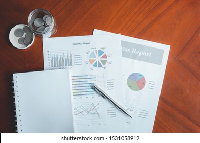 Notebooks, Money Jars With Coins And Summary Report With Bar Charts And Circles Placed On A Brown Wooden Table. Stock Analysis And Savings Concept. 