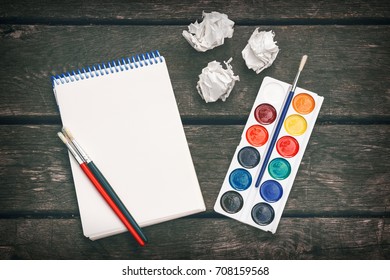 Notepad and blank sheet paper  colored paint  brushes   crumpled sheets paper the old wooden background  Workplace the artist  Never give up 

