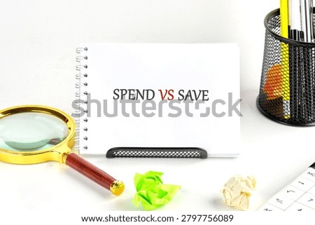 A notepad displays the words spend vs save. Nearby are a magnifying glass, crumpled papers, and office supplies on a desktop.