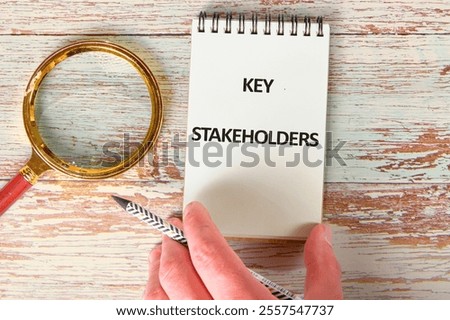 Notepad with the text Key Stakeholders displayed prominently, accompanied by a magnifying glass and a hand holding a pencil. This setup emphasizes the focus on crucial project contributors.