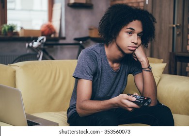 Nothing interesting to watch. Young African man holding remote control and looking bored while watching TV on the couch at home