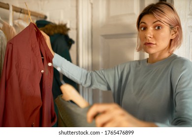 Nothing To Wear. Nervous Excited Young Female Going Out On First Date, Choosing Best Outfit, Having Worried Look, Holding Two Racks With Shirts. People, Lifestyle, Shopping And Clothing Concept