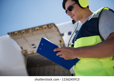Noting All Details. Low Angle Portrait Of Airport Worker Filling Out Documents. Open Cargo Door On Blurred Background