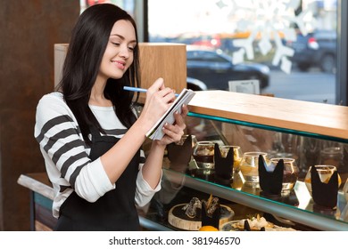 Noting carefully. Horizontal portrait of a gorgeous cheerful waitress writing notes working at the coffee shop - Powered by Shutterstock