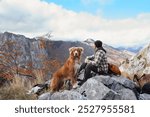 A Nova Scotia Duck Tolling Retriever stands with a hiker on a rocky mountain peak, surrounded by an autumn landscape. The rugged terrain and vast views add to the sense of adventure.