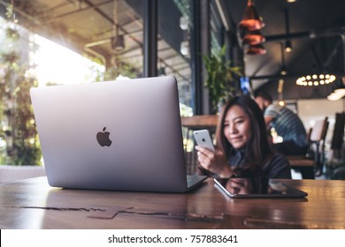 November 17th 2017 : A beautiful Asian business woman holding , using and looking at smart phone with laptop on wooden table in modern cafe , Chiangmai Thailand