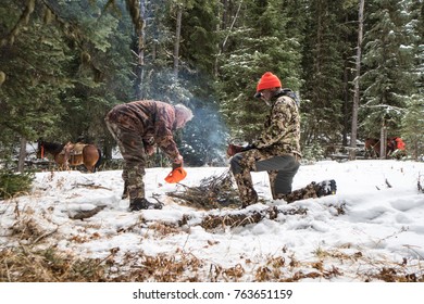 NOVEMBER 8 2017 - DILLON, MONTANA: Two Adult Male Hunters Try To Extinguish A Campfire In The Snow, Wearing Camouflage Hunting Gear At Their Deer Camp In The Winter